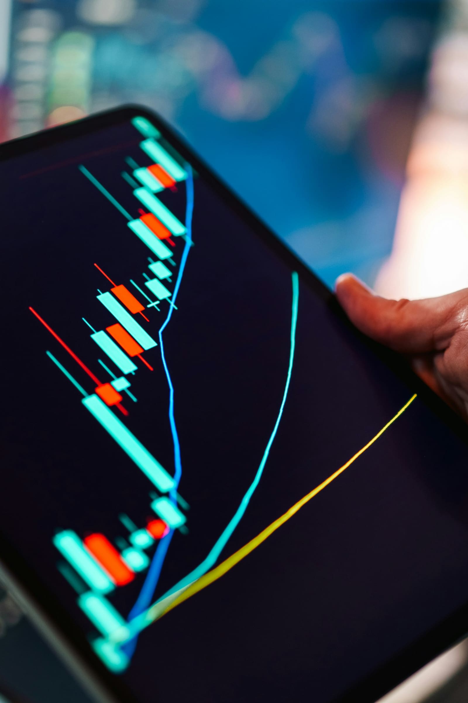 A hand holding a tablet showing a bright candlestick chart with two moving average lines running across it