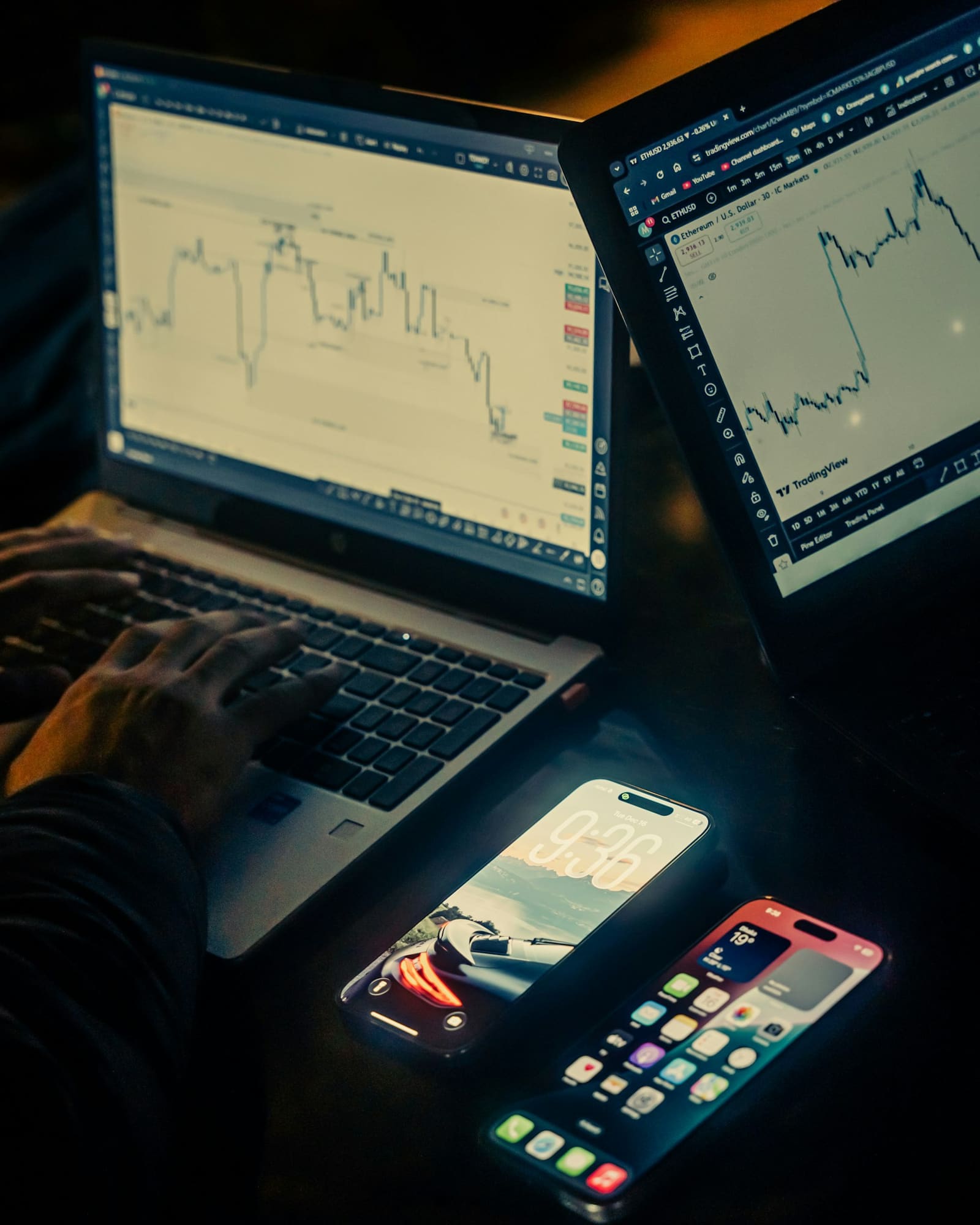 Two hands on a laptop keyboard with market charts open on the screens behind them